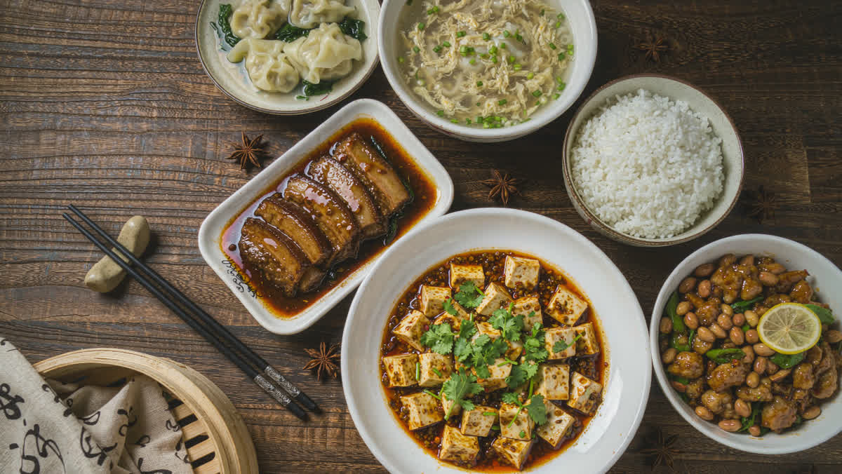 Overhead view of a Chinese table with mapo tofu, kung pao chicken, braised pork belly, steamed dumplings, scallion oil noodles and rice