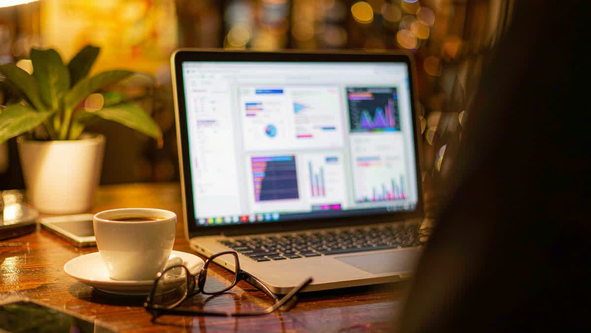 An open laptop on a wooden desk showing an abstract glowing chart dashboard, with a coffee cup and eyeglasses beside it.

