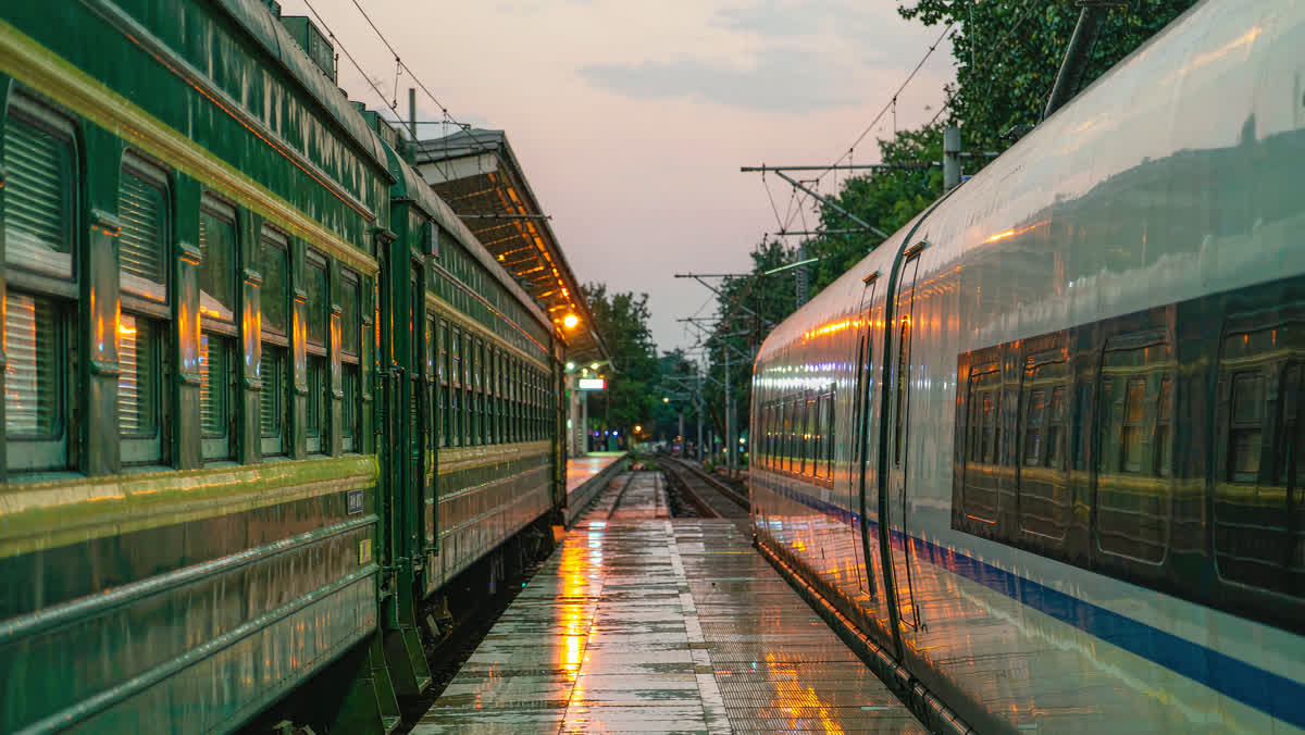 A white high-speed bullet train beside a green sleeper train on a platform at dusk