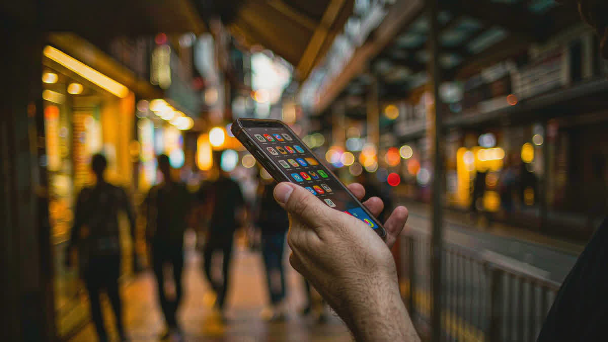 A traveler's hand holding a smartphone with a grid of colorful app icons, on a busy Chinese city sidewalk at dusk with blurred neon shopfronts.