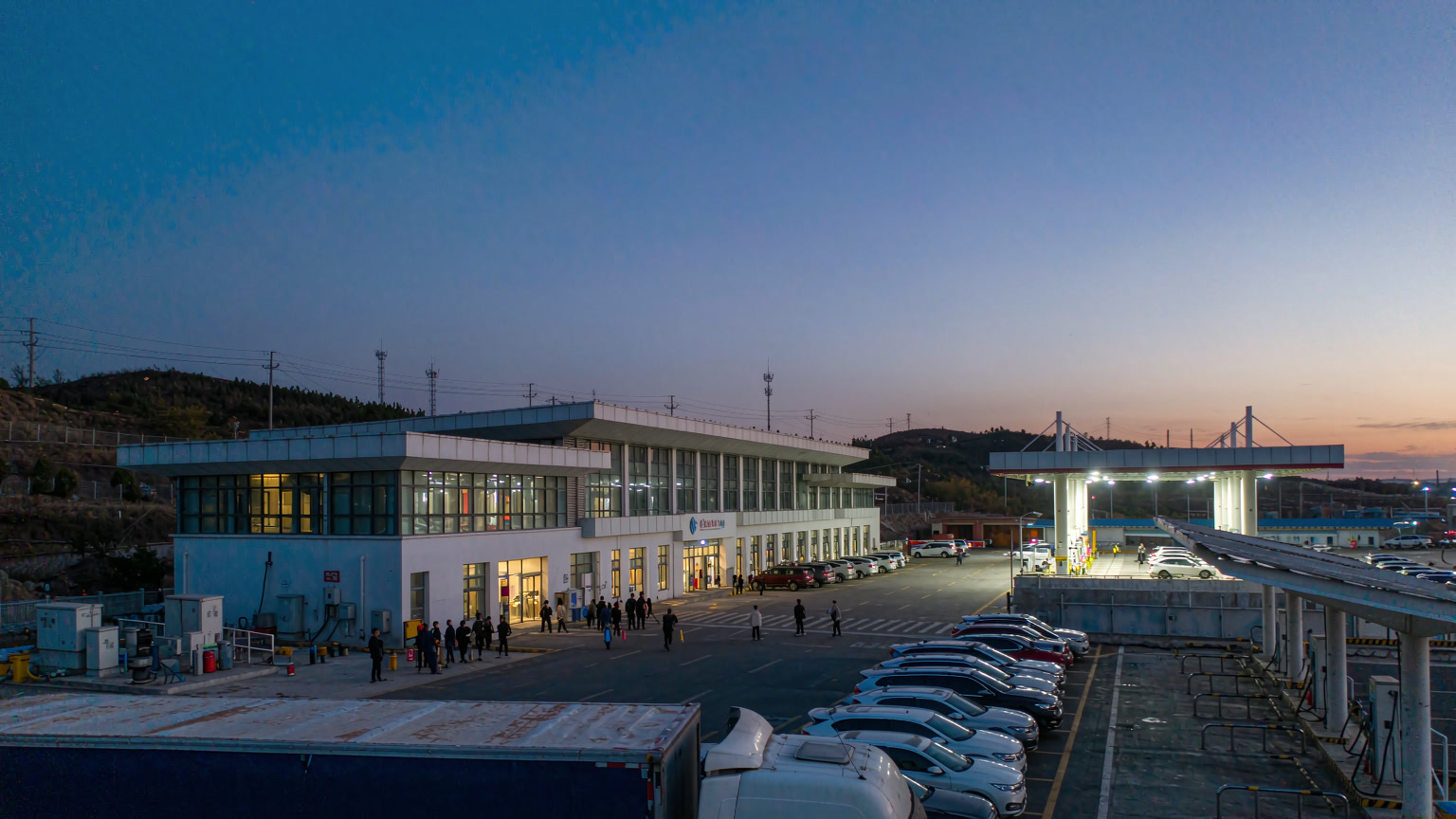 A Chinese expressway service area at dusk with parked cars, a long-haul truck, EV charging posts and a lit service building