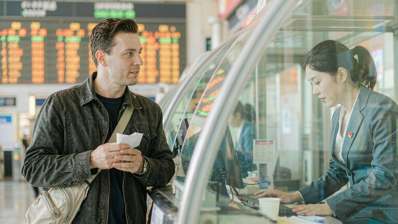 A foreign traveller holding a paper 客运记录 at a Chinese high-speed rail station ticket counter, waiting to be rebooked on the next train after overshooting his stop