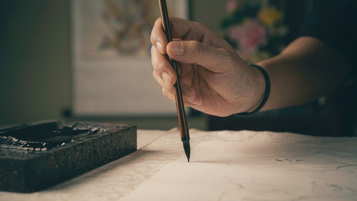 A calligrapher's hand holding a traditional brush over rice paper with an inkstone beside it