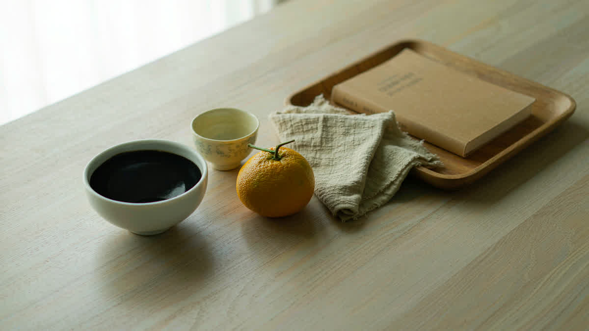 A still life of everyday objects on a light wooden table near a window: a porcelain bowl, a teacup, a mandarin orange and a closed book.
