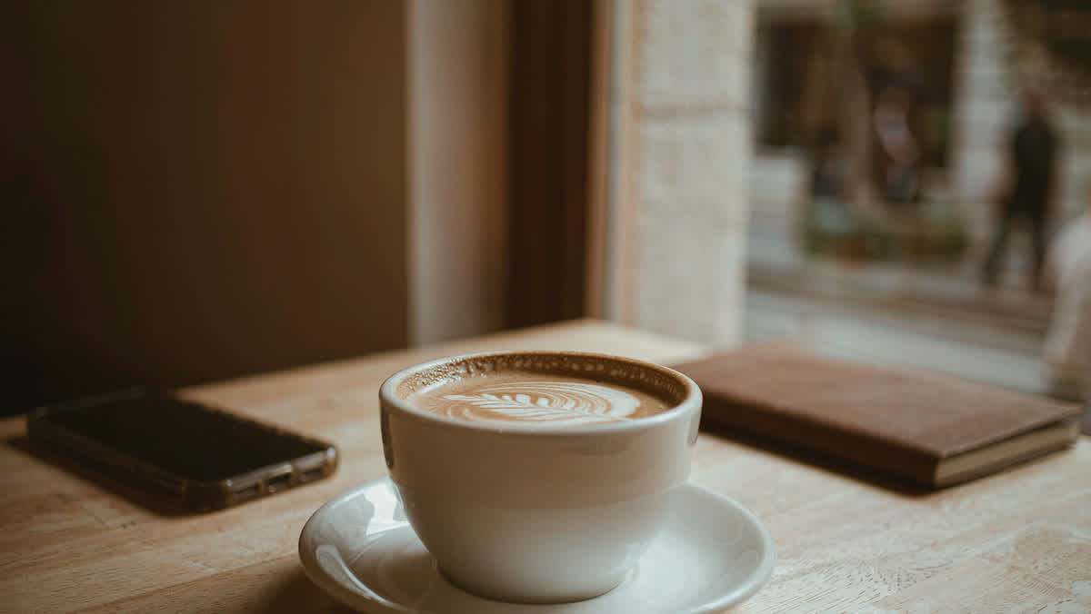 A latte with leaf latte art in a white ceramic cup on a light wooden cafe table near a large window.
