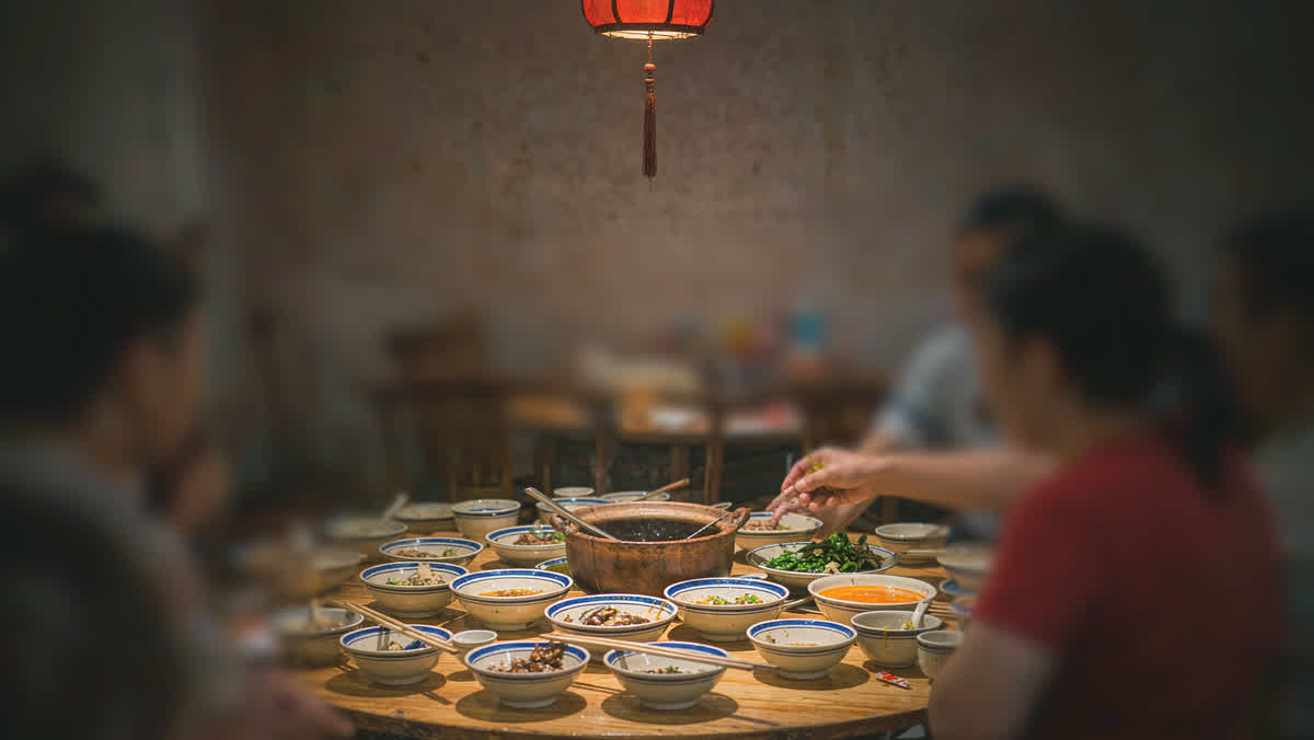 A round wooden dining table at a Chinese restaurant with small side dishes, a shared clay pot of soup and chopsticks on porcelain holders.

