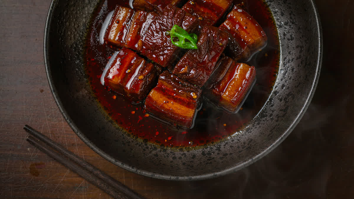 A traditional Chinese braised pork dish in a dark ceramic bowl on a wooden restaurant table with chopsticks beside it and gentle steam.