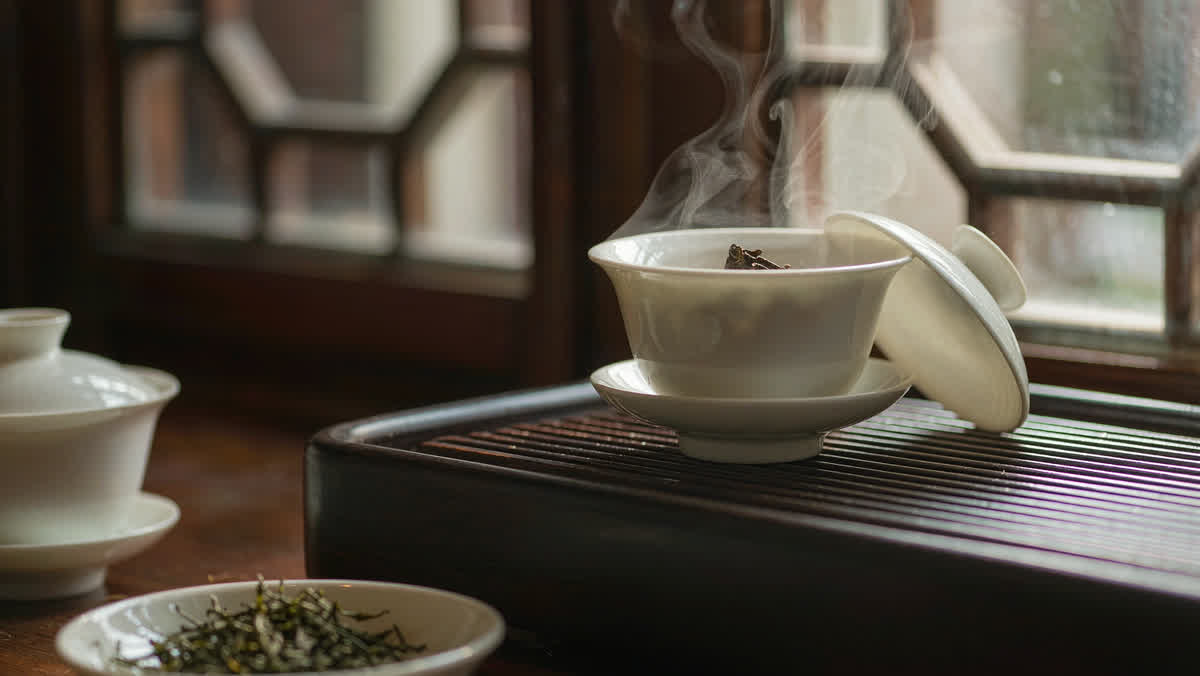 A white porcelain gaiwan with steam rising and dried tea leaves on a dark wooden tea tray
