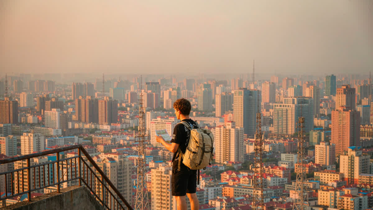 A young traveler with a canvas backpack seen from behind, gazing out at a wide modern Chinese city from a rooftop viewpoint.
