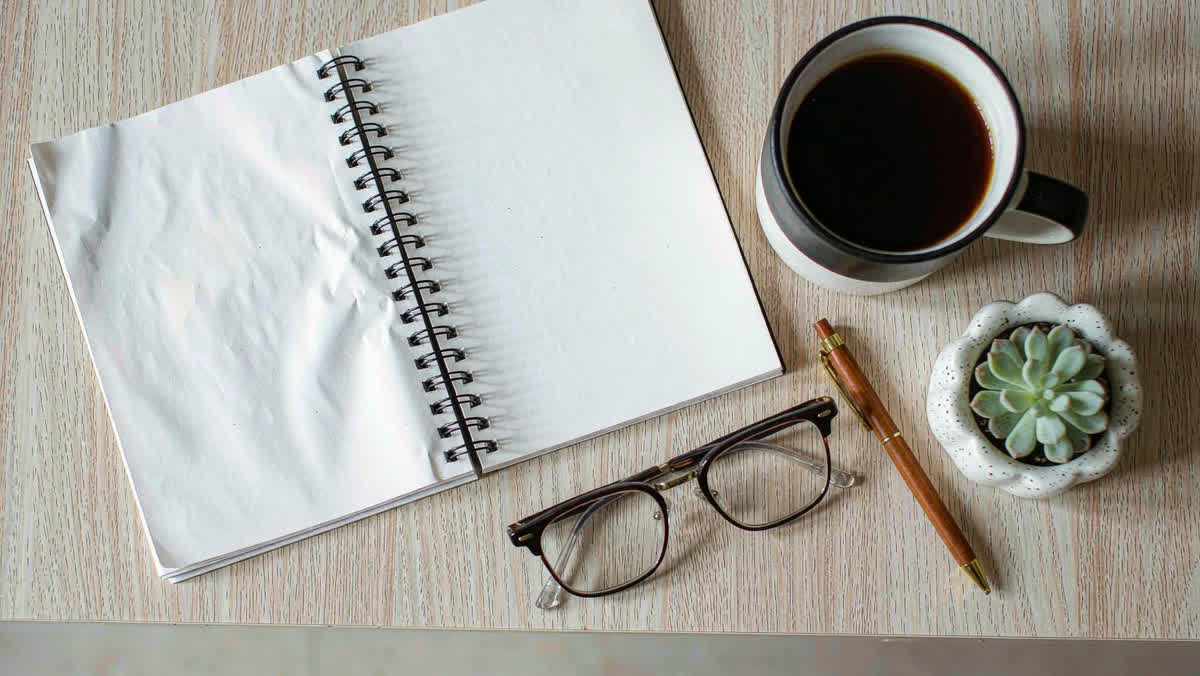 A top-down flat lay of an open ring-bound planner, a cup of coffee, a wooden pen and glasses on a light wood desk.
