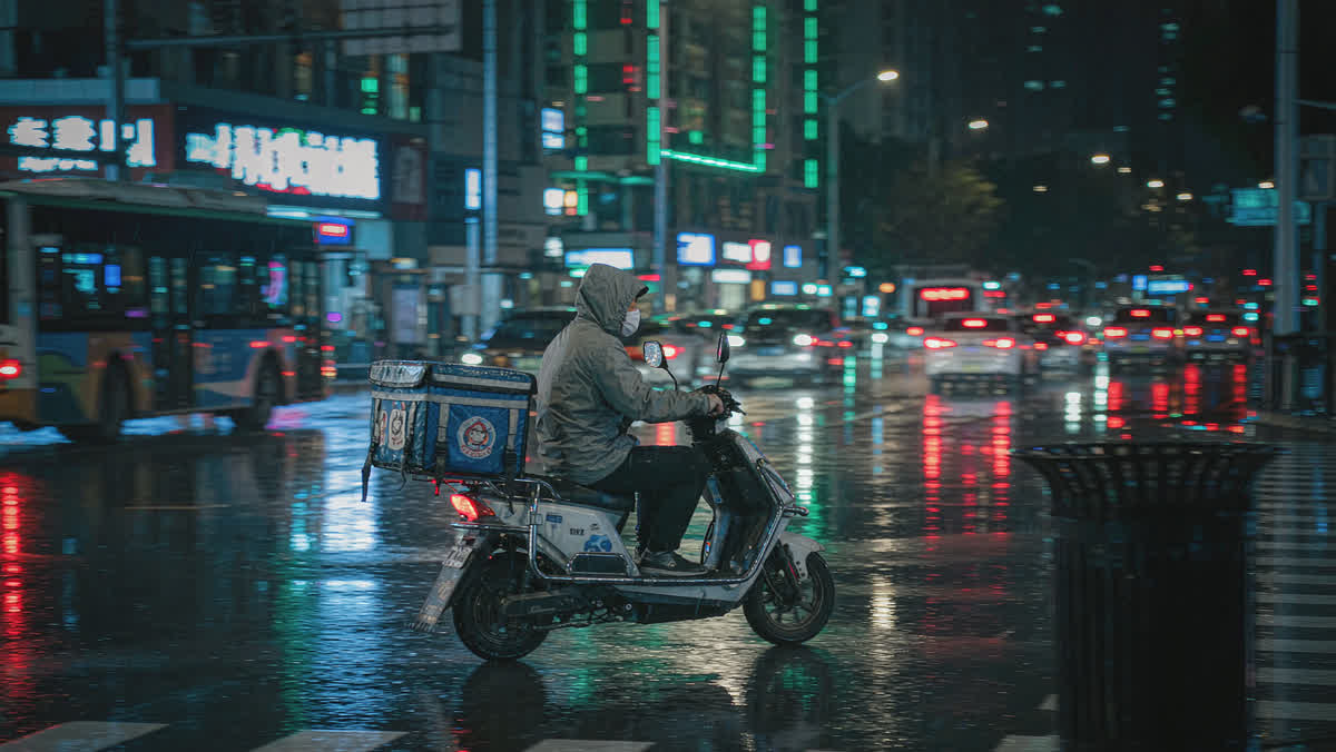 A food delivery rider on an electric scooter at a rainy night intersection in a Chinese city