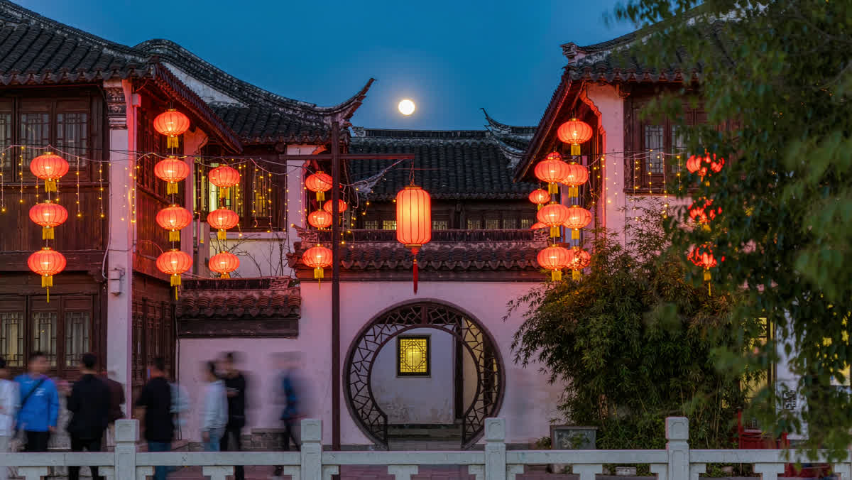A traditional Chinese old town square at night with glowing red lanterns strung between historic buildings and a softly blurred crowd.
