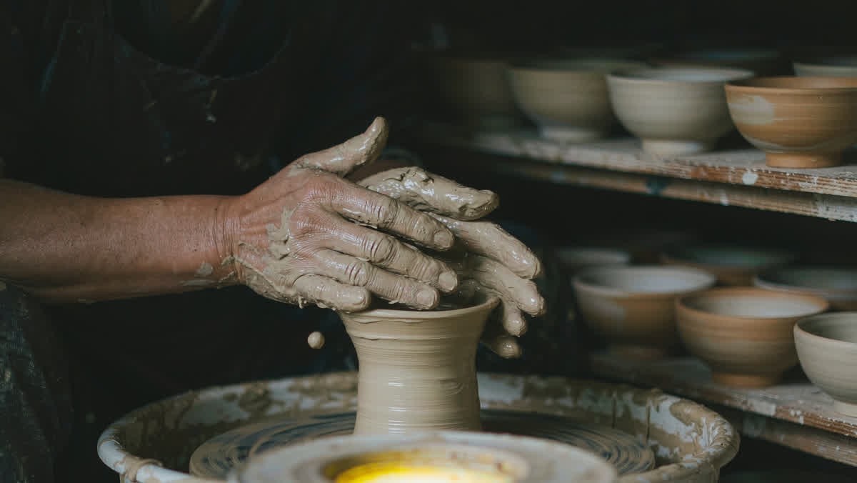 A potter's hands shaping wet clay on a spinning pottery wheel with shelves of bowls behind