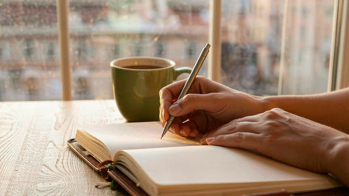 Hands writing in an open leather-bound journal on a bright wooden desk by a window in golden morning light.
