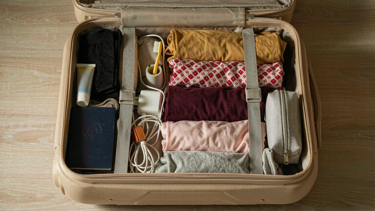 An open travel suitcase viewed from above, half-filled with folded clothes, a passport and a charger.
