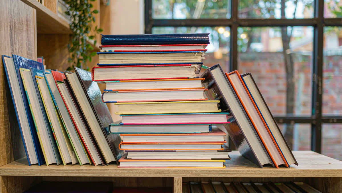 A neatly stacked pile of hardcover books with varied colored spines on a light wooden bookshelf.
