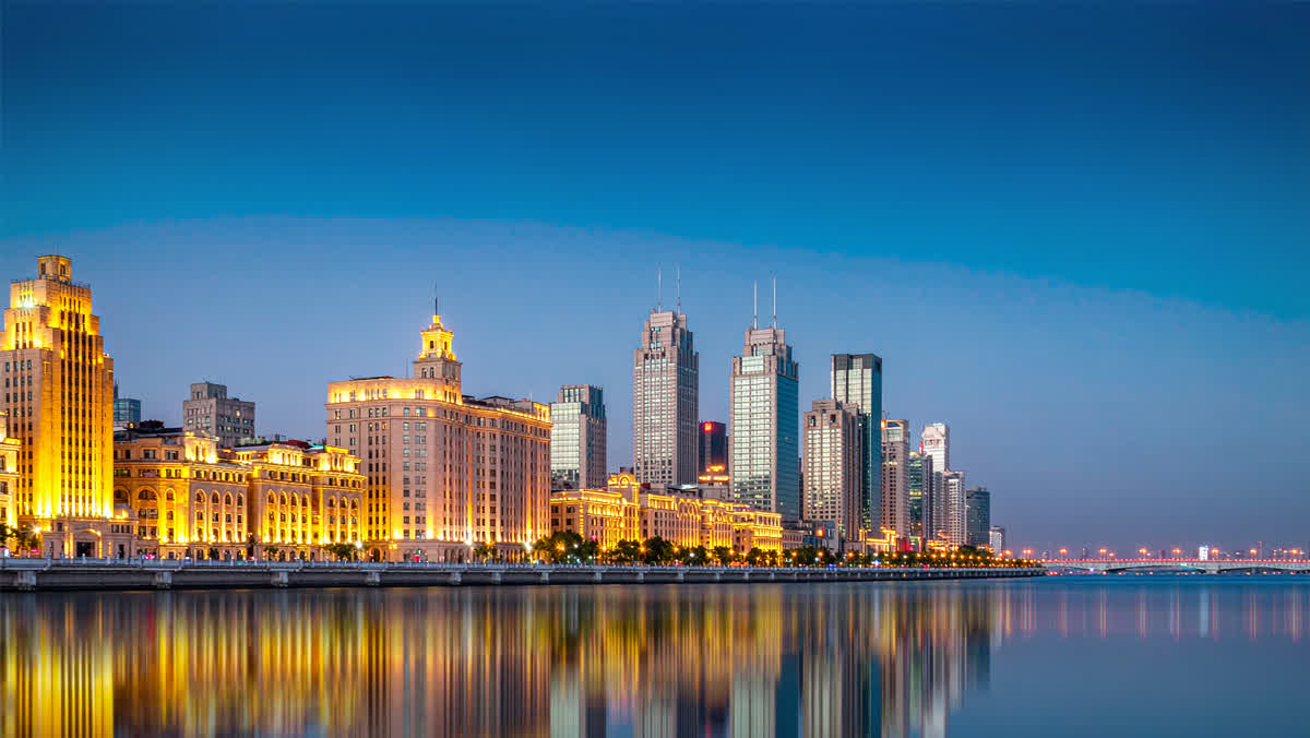 An illuminated riverside promenade of a major Chinese city at dusk with Art Deco and glass towers reflected in the water.
