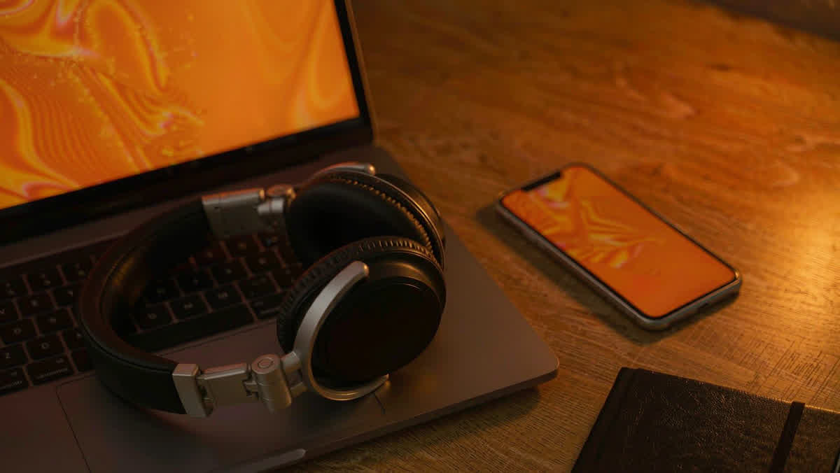 A laptop and a smartphone lying face-up side by side on a wooden desk, both screens softly glowing under a lamp.
