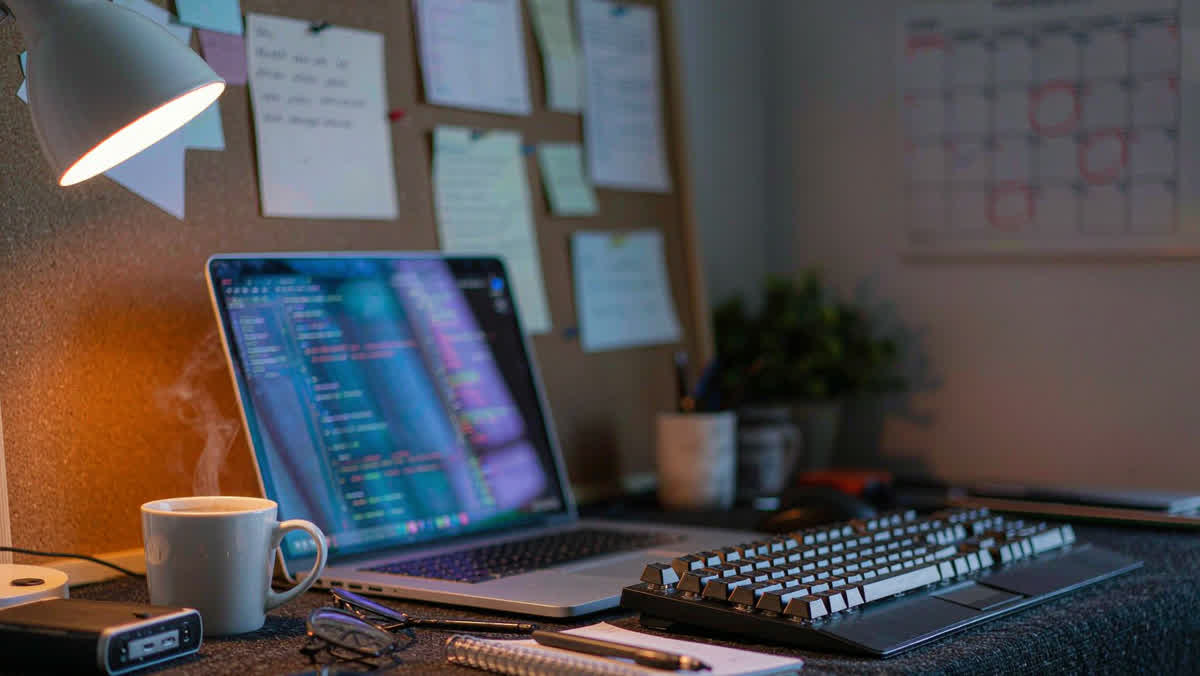 A laptop with glowing code on its screen next to a mechanical keyboard, a mug of coffee and a notebook under a warm desk lamp.
